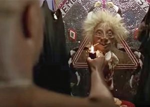 Screenshot from the movie "Major League". A fit, shirtless man holds the flame of a lighter to a cigar in the mouth of a short wild-haired voodoo loa figurine in a locker.