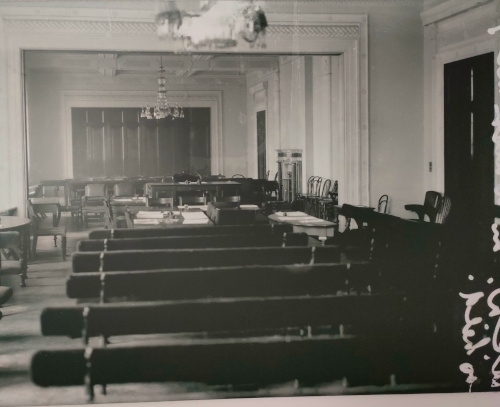 A black and white photo of a large room with molding on the walls and in archways. The room is full of benches and chairs, mostly visible from the back.