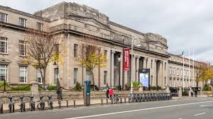 An image of a stone building, taken at a slight angle. It has columns and wings extending beyond a central point, and there are three visible floors.